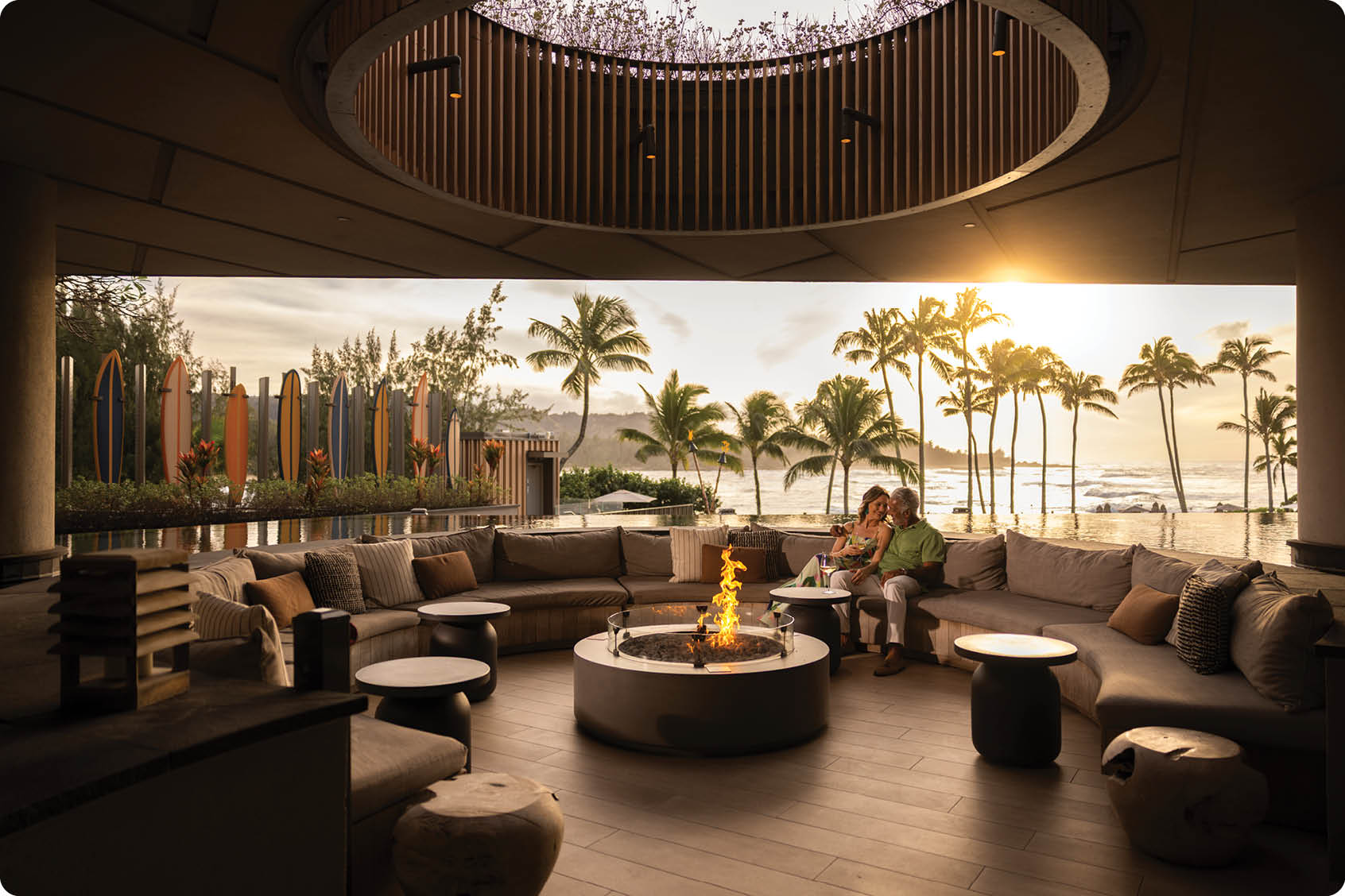 A man and woman are sitting on a patio overlooking the ocean. They are surrounded by chairs, a dining table, and a fire pit. The setting is tropical, with palm trees in the background. AI generated content