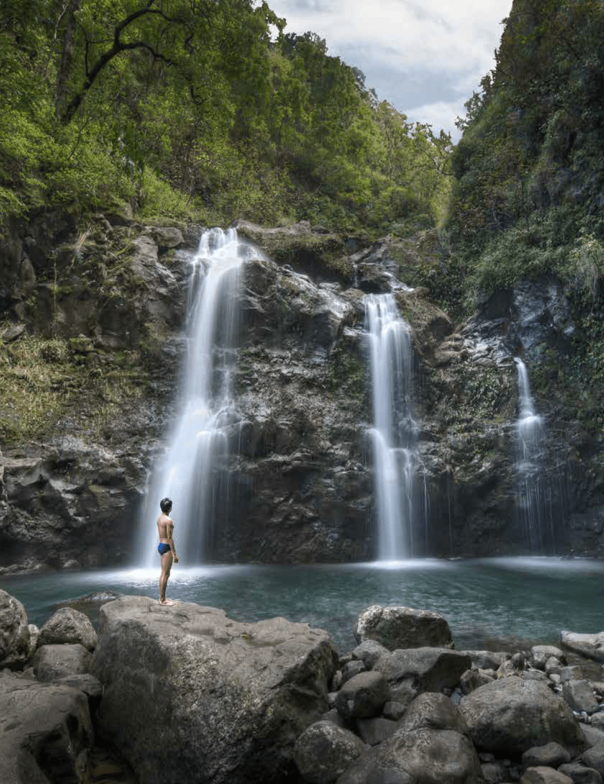 A woman stands on rocks near a waterfall, wearing a blue bikini. AI generated content