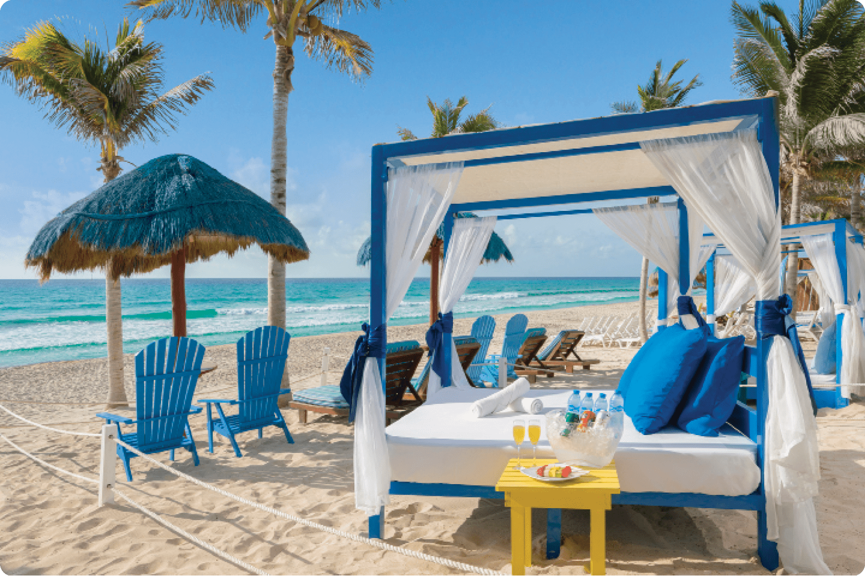 A beach scene with a blue canopy, a bed, and chairs. A table with a bowl of fruit and a bottle is also present. AI generated content